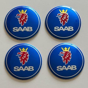 May include: Four circular emblems featuring the Saab logo. Each emblem has a deep blue background with a silver border. Centered is a red griffin head with a yellow crown, positioned above the silver text SAAB in a bold, sans-serif font.