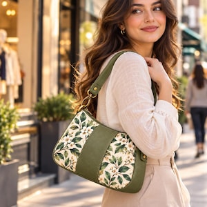 May include: A shoulder bag featuring a solid olive green center panel and side accents, contrasted with two floral patterned sections displaying white blossoms and green leaves. The bag includes a matching olive green shoulder strap with gold hardware.