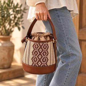 May include: A bucket bag featuring a brown leather base, handle, and trim, with a woven beige and brown geometric patterned body. The bag has a drawstring closure made of tan fabric, gold-toned metal hardware, and a structured cylindrical shape.