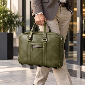 May include: A rectangular olive green leather briefcase featuring two top handles, a front zippered pocket with a dark brown zipper, and gold-toned metal hardware attachments for a shoulder strap on the sides. The bag has a smooth, structured finish.