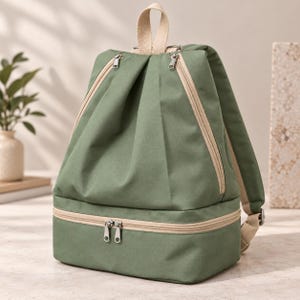 May include: A sage green backpack featuring a structured lower compartment with a horizontal zip, a main upper section with two diagonal zips, and beige fabric straps. The design includes a top handle and adjustable shoulder straps.