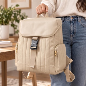 Könnte beinhalten: Ein beigefarbener, gesteppter Rucksack mit einem Überschlag und einer schwarzen Kunststoffschnalle, vertikalen Nähten sowie Seitentaschen mit goldfarbenen Reißverschlüssen. Der Rucksack verfügt über verstellbare Schultergurte und einen Tragegriff.