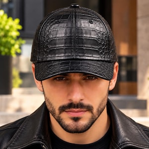 Puede incluir: Una gorra de béisbol negra con un patrón texturizado de piel de cocodrilo en toda la superficie. La gorra tiene una corona estructurada, una visera curva y un pequeño botón en la parte superior central. El material tiene un acabado brillante.
