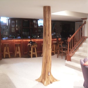 May include: A rustic basement bar with a large wooden tree trunk as a support beam. The bar has a wooden countertop and stools. There is a staircase leading up to an exit sign.