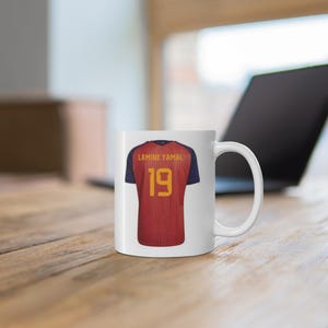 May include: A white ceramic mug featuring a graphic of a red and navy blue soccer jersey on the front. The jersey design includes the name LAMINE YAMAL and the number 19 printed in yellow text on the back of the shirt graphic.