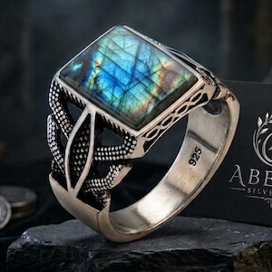 May include: A sterling silver ring featuring a square-cut labradorite gemstone with blue and gold iridescence. The band is engraved with intricate patterns and marked with 925. A black card with the silver text ABEERA SILVER ART is visible.