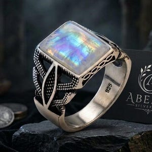 May include: A sterling silver ring featuring a rectangular iridescent moonstone cabochon set in a decorative band with intricate cut-out patterns. The inner band is stamped with 925. A black card displays the silver logo text ABEERA SILVER ART.