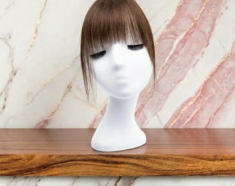 Op de afbeelding: Een bruin haartopstuk met rechte, dunne pony op een wit etalagehoofd. Het haar is glad met een natuurlijk ogende scheiding en lokken die het gezicht omlijsten, ontworpen om extra volume en dekking aan het kapsel toe te voegen.
