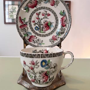 May include: A white porcelain teacup and saucer set featuring a hand-painted floral pattern with pink, blue, and green accents. Both pieces are decorated with a black geometric Greek key border design along the rims and center.