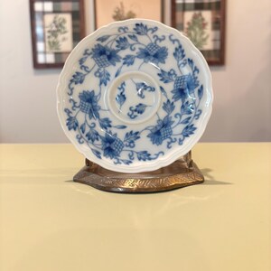May include: A white porcelain saucer featuring a scalloped edge and a blue floral pattern with intricate vines and blossoms. The saucer is displayed upright on a metallic stand, showcasing a central circular indentation and detailed blue motifs.