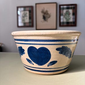 May include: A beige ceramic bowl with a glossy finish, featuring a hand-painted dark blue heart motif at the centre, flanked by stylised leaf patterns, and accented with horizontal blue bands encircling the upper and lower sections of the bowl.