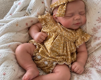 May include: A realistic baby doll with closed eyes, wearing a mustard yellow floral romper with ruffled sleeves and white snap buttons, paired with a matching mustard yellow floral bow headband, resting on a white muslin blanket with a floral print.