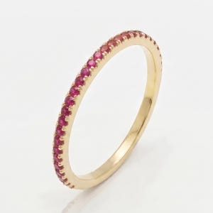 Puede incluir: Un anillo fino de oro amarillo con una hilera continua de pequeños rubíes redondos de color rojo vibrante engastados en el borde exterior. El metal tiene un acabado pulido y reflectante, y las gemas son de tamaño uniforme y circular.