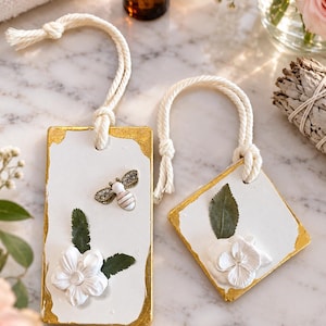 Puede incluir: Dos adornos de arcilla blanca con bordes pintados en oro y colgadores de cuerda color crema. El adorno rectangular tiene un dije de abeja y una flor blanca con hojas verdes. El adorno cuadrado presenta una flor blanca con hojas verdes.