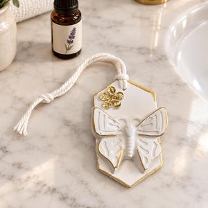 May include: A white ceramic hanging ornament in a hexagonal shape featuring a raised butterfly relief and a small gold flower accent. The edges are trimmed in gold, and it includes a thick white cotton cord for hanging. A small amber glass bottle.