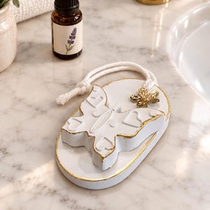 May include: An oval white ceramic plaque featuring a raised butterfly design with heart-shaped cutouts and gold-painted edges. A white braided rope is attached to the top, and a small gold-toned metallic butterfly charm is secured to the rope.