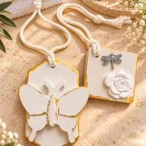 Puede incluir: Dos adornos colgantes blancos con bordes pintados en dorado. Uno tiene forma hexagonal con un relieve de mariposa, y el otro es cuadrado, decorado con un pequeño dije de libélula plateada y un relieve de flor blanca esculpida.
