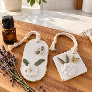 Puede incluir: Dos adornos colgantes blancos, uno ovalado y otro cuadrado, con flores blancas en relieve, hojas verdes y pequeños detalles metálicos de abeja y mariposa. Cada adorno incluye un lazo de cuerda de color crema para colgar y bordes dorados.