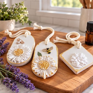 Puede incluir: Tres adornos colgantes blancos con bordes pintados en oro. Dos son de forma ovalada con relieves de margaritas blancas y doradas y abejas. El tercero es un adorno cuadrado con un relieve floral blanco. Cada uno tiene un lazo de cuerda.