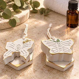 May include: Two white butterfly-shaped hanging ornaments with gold-painted edges. Each ornament features a textured butterfly design on a hexagonal base and includes a white string loop for hanging. The surfaces are matte white with gold accents.