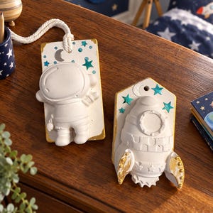 May include: Two white wall-mounted plaques featuring space-themed designs. One plaque depicts a 3D astronaut figure, while the other shows a rocket ship. Both plaques are decorated with teal painted stars and a silver crescent moon accent.