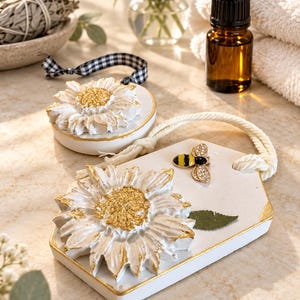 Puede incluir: Dos adornos de arcilla blanca con relieves de girasol pintados en dorado. Uno es circular con una cinta de cuadros negros y blancos, y el otro es hexagonal con un dije de abeja, un detalle de hoja verde y una cuerda blanca gruesa.