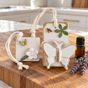 May include: Two white hanging air freshener plaques with gold-painted edges. One square plaque features a gold butterfly and a white flower. The rectangular plaque features a gold bee, a green leaf, and a large white butterfly. Both have rope loops.