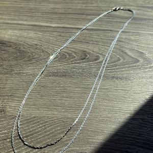 May include: A delicate silver-toned chain necklace featuring a double-layered design with fine, interlocking links. The metallic surface reflects light, creating a bright, polished appearance along the length of the thin, flexible metal strands.