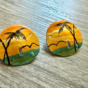 May include: A pair of round sand dollar ornaments featuring a hand-painted tropical beach scene. The design includes a vibrant orange sky, a green ocean horizon, black silhouettes of palm trees, and small birds against the sunset background.