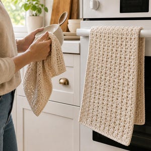 Puede incluir: Un paño de cocina rectangular de color crema con un patrón de ganchillo texturizado similar a un gofre. El paño está colgado del asa de un horno blanco y también se muestra siendo sostenido, destacando la construcción de hilo grueso.