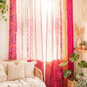 Peut inclure: Une paire de rideaux pleine longueur présentant un motif patchwork de tissus rose, orange et rouge vifs avec divers motifs floraux et géométriques. Les rideaux sont suspendus à une tringle en bois par des pattes et laissent passer la lumière.