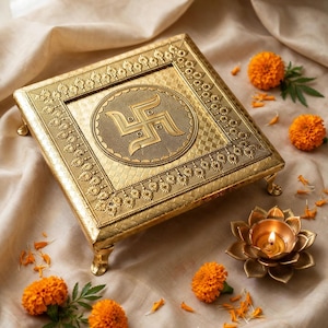 May include: A square, gold-colored metal pedestal with four short legs, featuring an embossed swastika symbol in the center surrounded by intricate geometric and floral patterns on the border, accompanied by a lotus-shaped metal oil lamp.