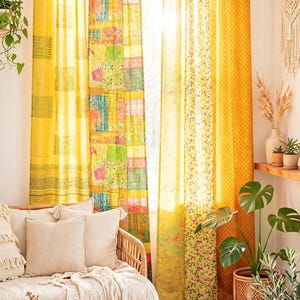 Op de afbeelding: Een set kamerhoge gordijnen met een levendig patchworkontwerp in tinten geel, groen en oranje. De gordijnen hangen aan een houten roede met stoffen lussen en tonen een mix van bloemrijke, geometrische en effen patronen.