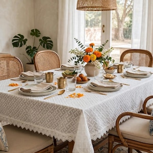 Peut inclure: Une nappe rectangulaire blanche avec un motif géométrique en treillis et une bordure en dentelle festonnée, recouvrant une table à manger dressée avec des assiettes en céramique, des verres transparents, des tasses en laiton et un vase central.