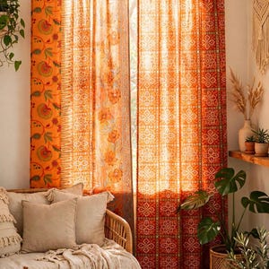Puede incluir: Un par de cortinas de cuerpo entero con una vibrante paleta de colores naranja y rojo con intrincados patrones florales y geométricos. Las cortinas están suspendidas de una barra de madera mediante pestañas de tela y permiten que la luz se filtre.