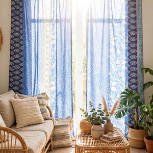 Op de afbeelding: Een paar kamerhoge gordijnen met een patchworkontwerp in diverse blauwe en witte tinten met bloemen- en paisleymotieven. De gordijnen hangen aan een houten roede met stoffen lussen en zijn semi-transparant, waardoor het zonlicht erdoorheen valt.