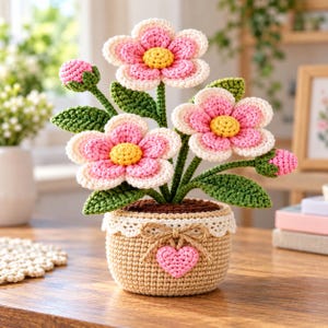May include: A crocheted flower arrangement featuring three pink and white blossoms with yellow centres and two small pink buds. The flowers have green stems and leaves, all contained in a tan crocheted pot decorated with a white lace trim and a pink heart.