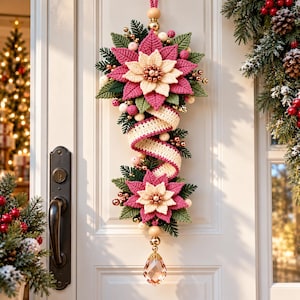 May include: A vertical hanging door decoration featuring two crocheted poinsettia flowers in pink and cream, green crocheted leaves, a cream crocheted ribbon, small metallic gold and pink spheres, and a clear faceted crystal drop at the base.
