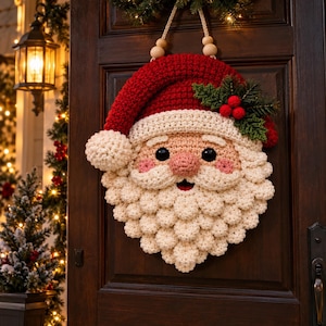 Puede incluir: Un adorno de pared de Papá Noel tejido a ganchillo con un gorro rojo con pompón blanco, una barba blanca texturizada, nariz rosa, ojos negros y una rama de acebo verde con tres bayas rojas unida al borde del gorro.