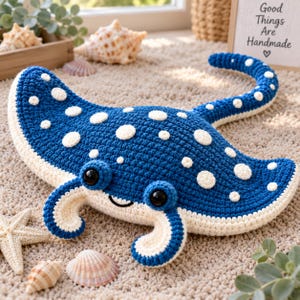 May include: A crocheted stingray plush toy featuring a deep blue body with white polka dots, a cream-colored underside, and large black plastic eyes. The stingray has a long, slender tail and curved pectoral fins, crafted with visible yarn stitches.