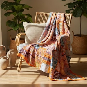 May include: A rectangular throw blanket featuring a patchwork design of various geometric and floral patterns in shades of orange, blue, and white. The blanket is draped over a light-colored wooden chair with a beige cushion, showcasing its texture.