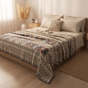 Könnte beinhalten: Eine gewebte Wohndecke mit einem sich wiederholenden geometrischen Muster in Creme, Dunkelblau und Rostorange. Die Decke hat einen Fransenrand und liegt auf einem Bett mit beigen Kissen, darunter ein quadratisches Kissen mit Strickstruktur.