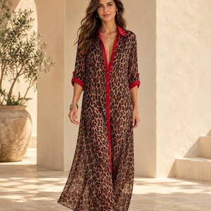 May include: A floor-length button-down shirt dress featuring a brown and black leopard print pattern. The garment includes a solid red collar, a red button placket, and red cuffs on the three-quarter length sleeves. The fabric is sheer and lightweight.
