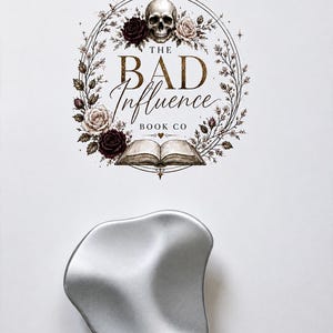 May include: A silver-coloured, organic-shaped metal tin with a smooth, matte finish. Above the tin is a circular logo featuring a human skull, dark and light roses, and an open book, with the text THE BAD INFLUENCE BOOK CO in the centre.