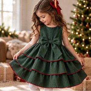 May include: A sleeveless dark green dress featuring a large bow at the waist and a multi-tiered skirt with red trim along the edges. The dress has a round neckline and a full, flared silhouette. A red hair bow is worn in the long wavy hair.
