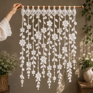 May include: A white crochet wall hanging featuring multiple vertical strands of interconnected floral and leaf motifs suspended from a wooden rod. The intricate lace-like design consists of repeating five-petaled flowers and small leaves.