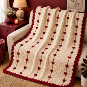 May include: A cream-colored crocheted throw blanket featuring vertical rows of small, dark red hearts. The blanket is finished with a scalloped dark red border along all edges, creating a textured, repeating pattern across the entire surface.