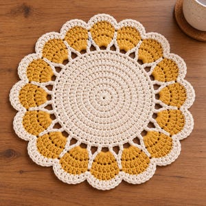 May include: A round, crocheted doily featuring a central cream-colored circular pattern surrounded by a scalloped border of mustard yellow and cream-colored yarn. The intricate design consists of dense, textured stitches forming a flower-like shape.