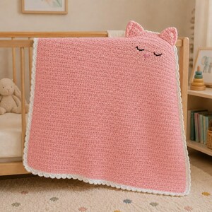 May include: A square, pink crocheted blanket featuring a cat-themed design at the top with two small ears, embroidered closed eyes, and a pink nose. The blanket is finished with a white scalloped crochet border along all four edges.
