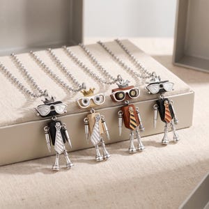 May include: Four robot-shaped pendants on silver-toned ball chains. Each robot features a crown, glasses, a rectangular torso with a striped tie, and articulated limbs. The robots vary in color, including black, light wood, dark wood, and black finishes.